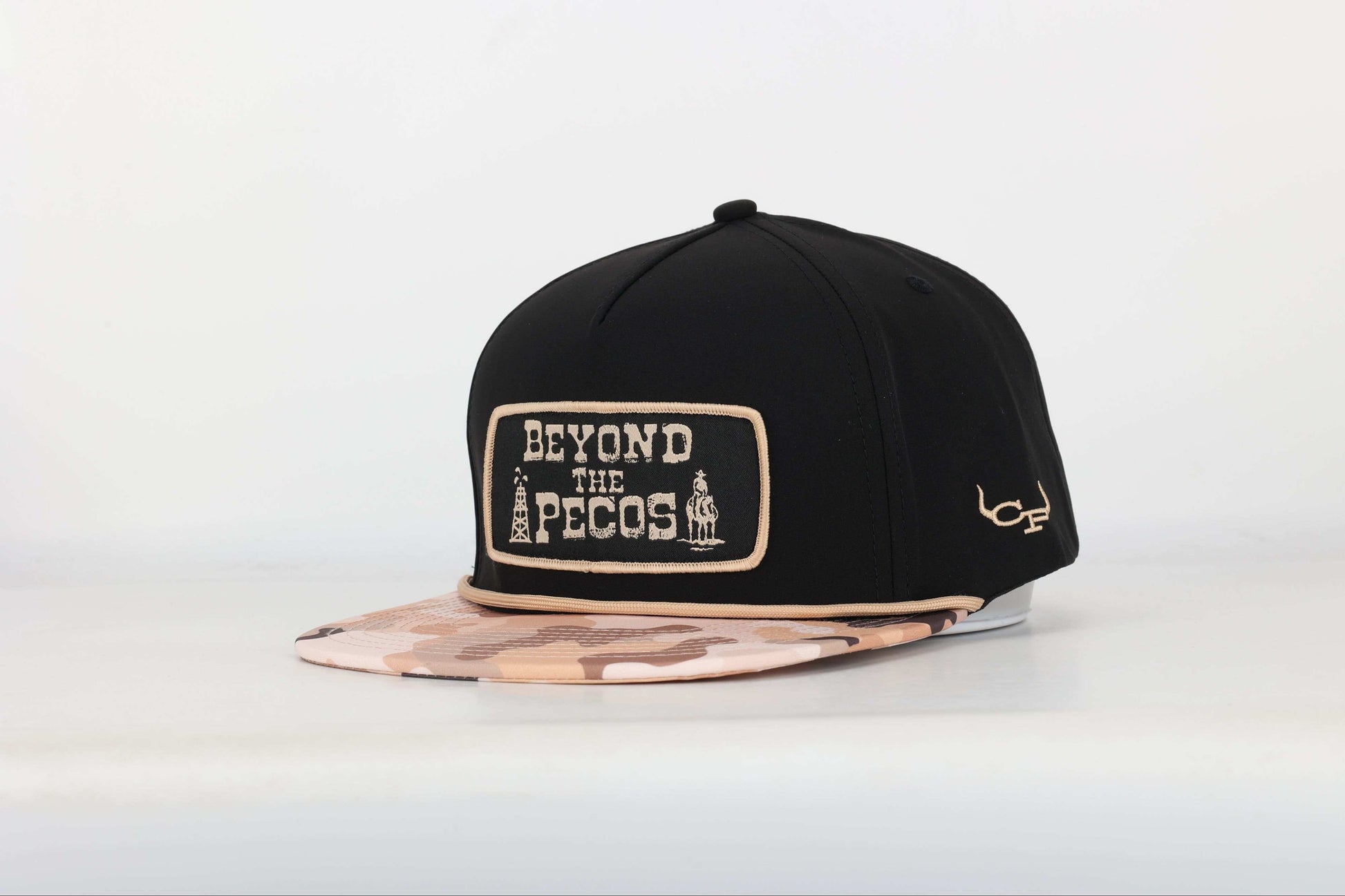Black snapback cap with "Beyond the Pecos" woven patch on the front in old gold, featuring an oil rig and cowboy graphic, old gold rope, and a desert camo print bill. Angled View