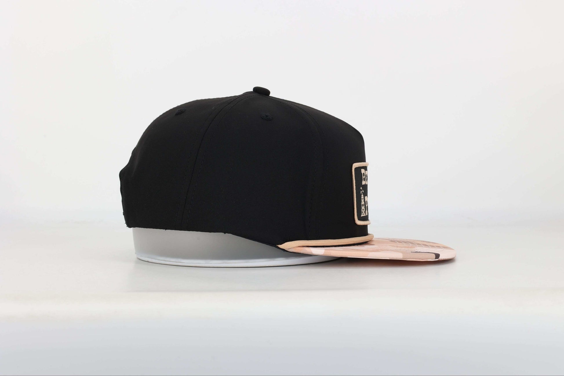 Black snapback cap with "Beyond the Pecos" woven patch on the front in old gold, featuring an oil rig and cowboy graphic, old gold rope, and a desert camo print bill. Left Side View