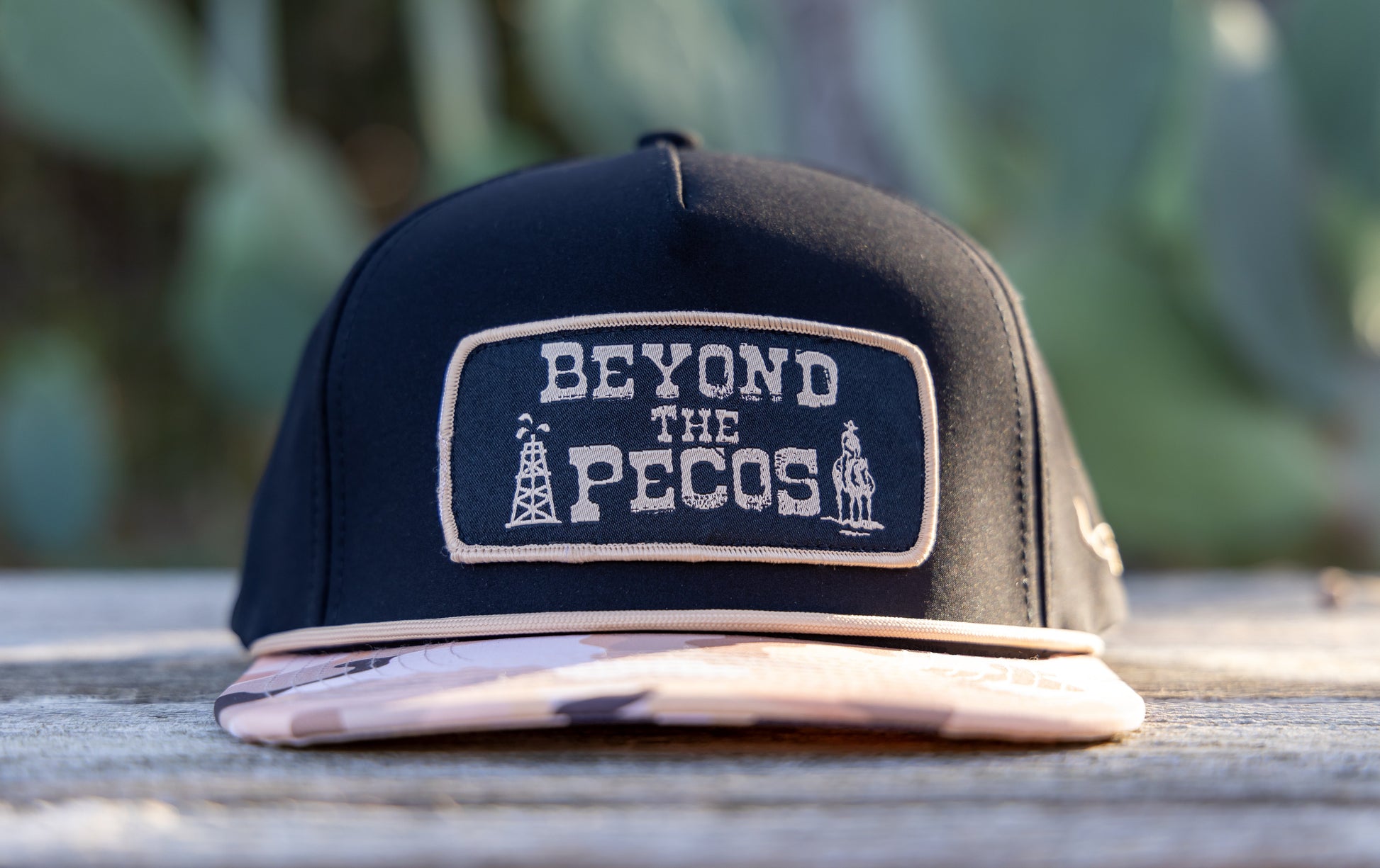Black snapback cap with "Beyond the Pecos" woven patch on the front in old gold, featuring an oil rig and cowboy graphic, old gold rope, and a desert camo print bill. Front View. Cactus in background. Product shot