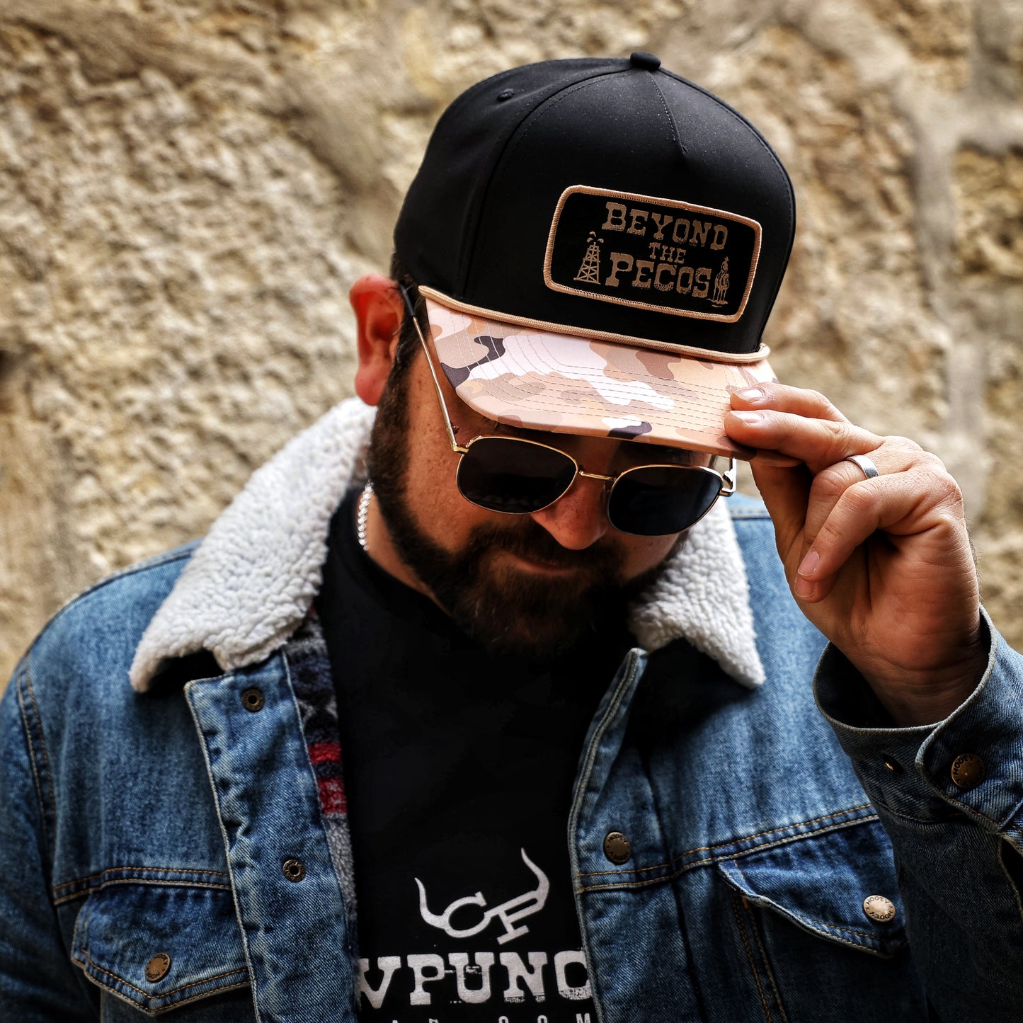 Black snapback cap with "Beyond the Pecos" woven patch on the front in old gold, featuring an oil rig and cowboy graphic, old gold rope, and a desert camo print bill. Male lifestyle model