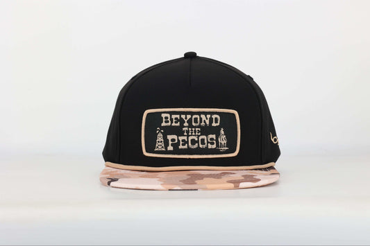 Black snapback cap with "Beyond the Pecos" woven patch on the front in old gold, featuring an oil rig and cowboy graphic, old gold rope, and a desert camo print bill. Front View