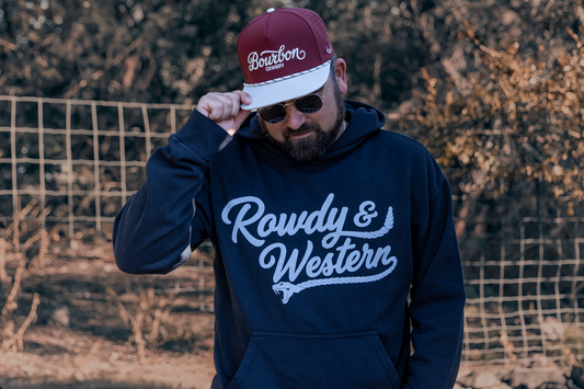 A man wearing a Black hoodie with 'Rowdy & Western' in white script on the front, a matching maroon and white baseball cap, and sunglasses, standing outdoors in front of a wire fence and dry brush.