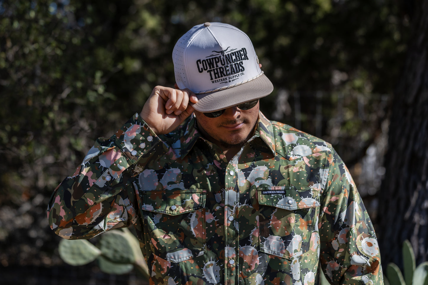 Man wearing a floral camo pearlsnap shirt with a pattern of prickly pear cactus 'Texas Regions Pearlsnaps Collection' branding and a 'Cowpuncher Threads' white cap.