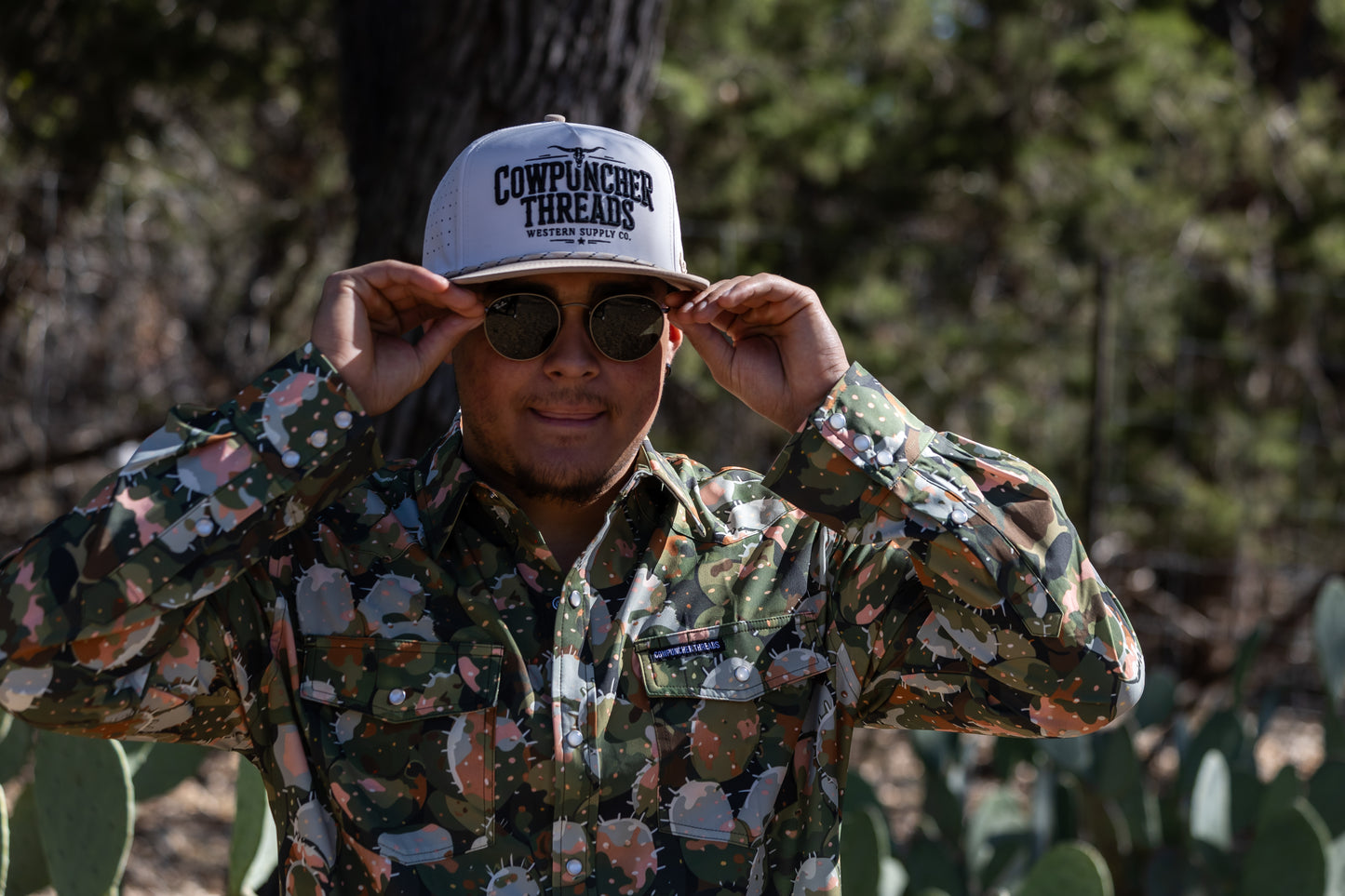Person wearing a Cactus camouflage shirt and white 'Cowpuncher Threads' cap in a natural setting with trees and prickly pear cacti.