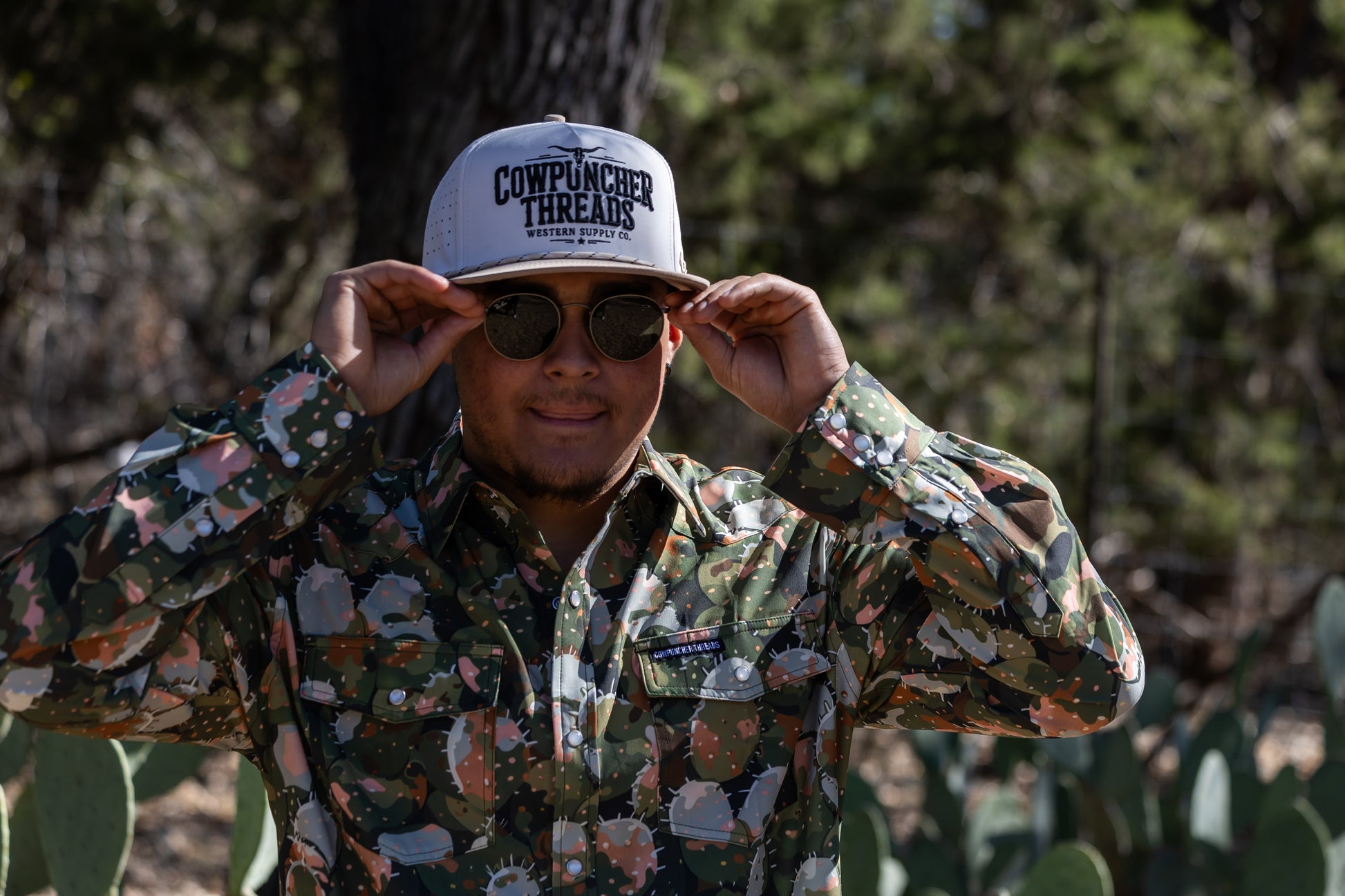 Person wearing a Cactus camouflage shirt and white 'Cowpuncher Threads' cap in a natural setting with trees and prickly pear cacti.