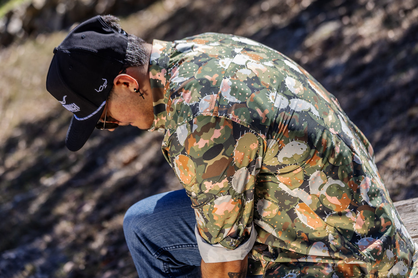 Person wearing a prickly pear cactus camouflage pearlsnap shirt. and black CP cap outdoors.