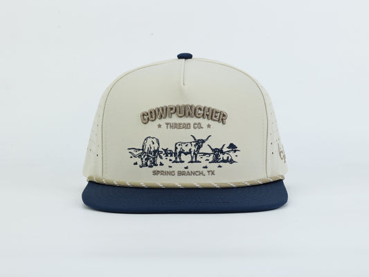 A cream-colored snapback cap with a navy blue flat brim and matching navy button on top. The front panel features a dark navy graphic of longhorn cattle grazing with the text 'COWPUNCHER THREAD CO."