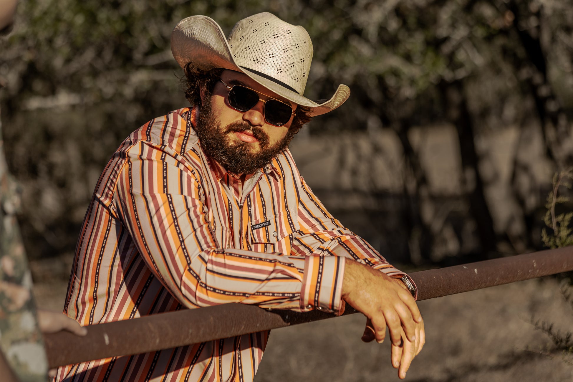 Man wearing a orange and purple stripped pearlsnap and cowboy hat and sunglasses leaning against a railing with a natural background