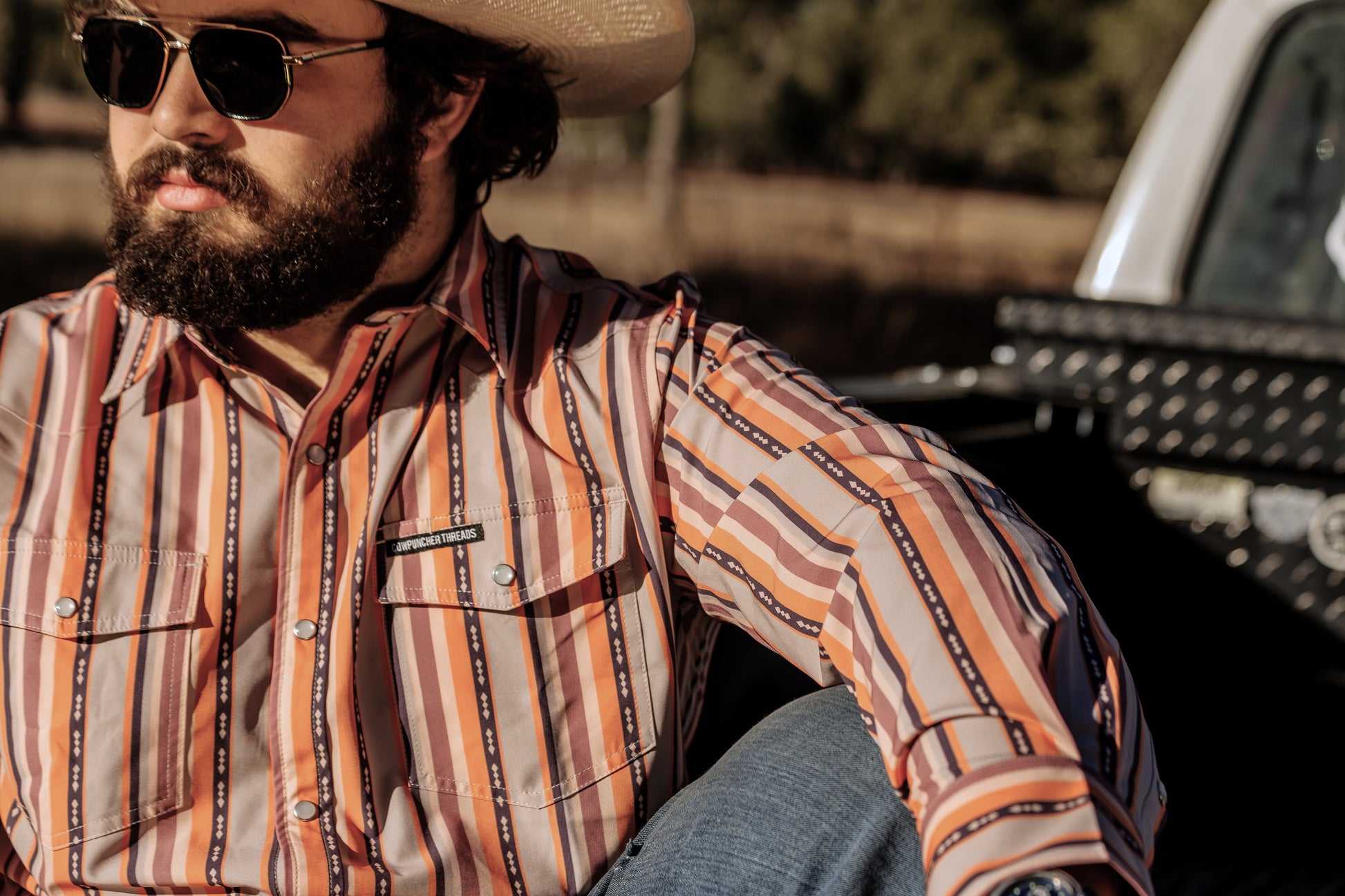 Man wearing a orange and purple striped pearlsnap shirt and cowboy hat sitting on the tailgate of a white ford truck.